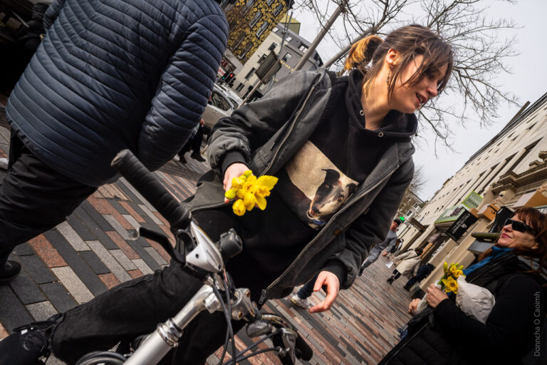 A woman on a bike with daffodils in her hand talks to someone out of the frame. Another woman to the right also has the same flowers in her hand.