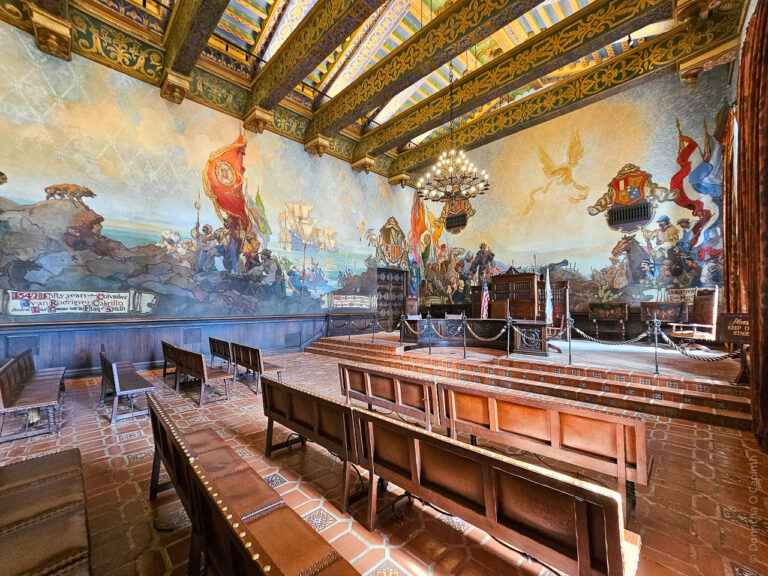 A view of the mural room, with seating in front, and a stage with furniture. The murals adorn the walls showing the sometimes inaccurate history of the state.