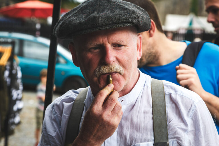 An older man wearing a flat cap and a white shirt with suspenders. He has a mustache and is holding a bullet to his lips. The background is slightly blurred, with other people and a car visible. The setting appears to be outdoors, possibly at a market or public event.