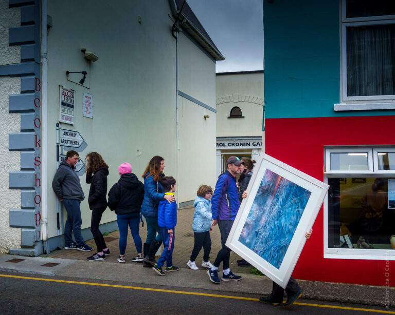 A man carries a painting of the sea with visible waves crossing the water. He's walking along a road in Dingle while people walk towards him.