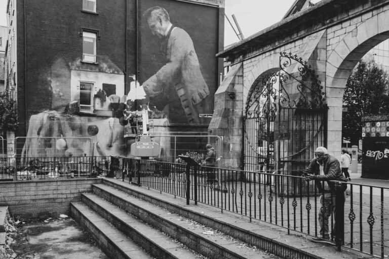 A black and white photograph depicting Bishop Lucey Park. A large mural on the side of a building features a man in a suit, appearing to be painting or working on something. In front of the mural, a person is elevated on a platform, possibly working on the mural itself. The scene includes an arched stone gate with ornate ironwork. Two people are visible: one leaning against a railing and another standing nearby. The setting appears urban, with steps leading up to the area where the people are located.