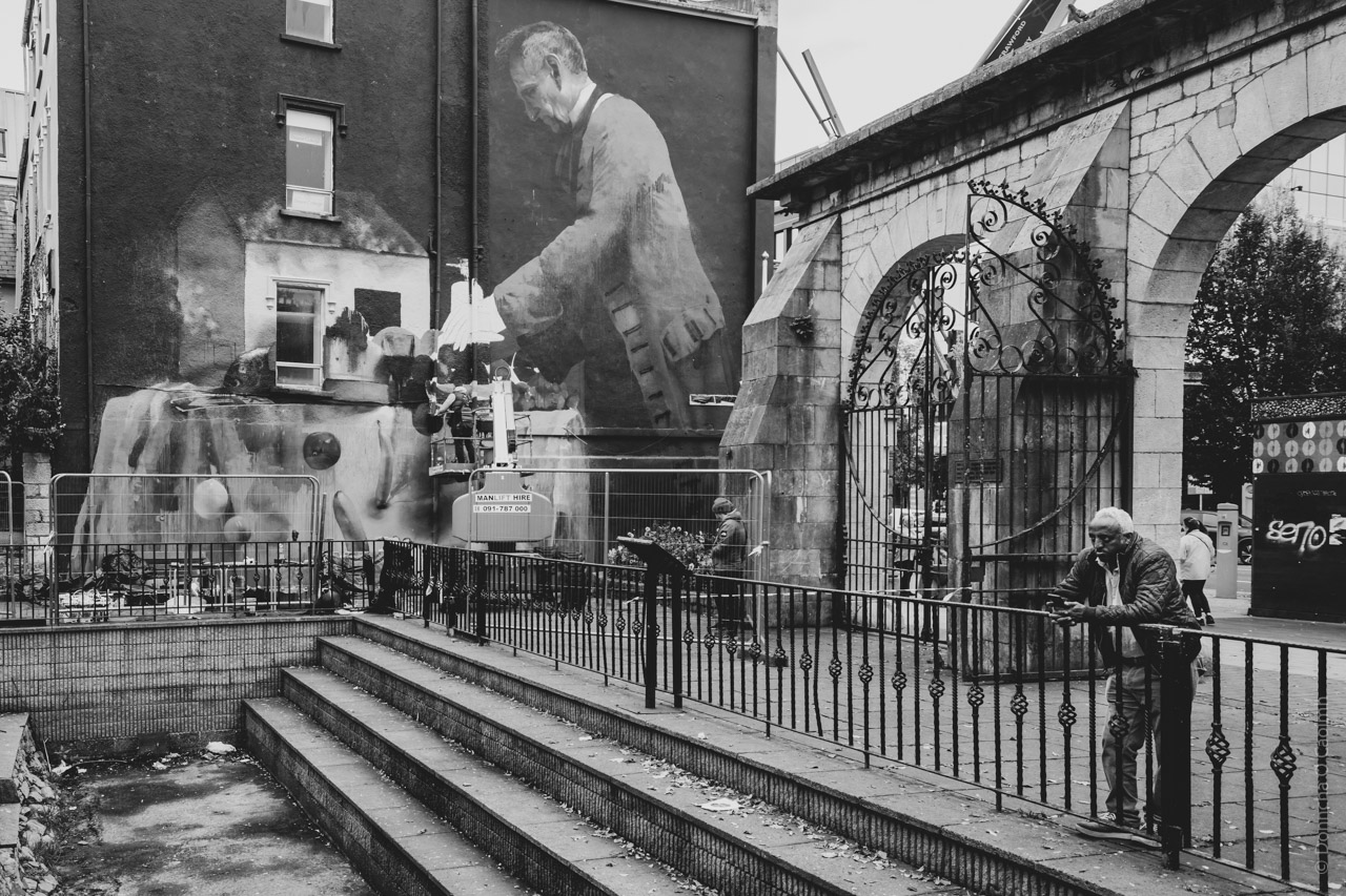 A black and white photograph depicting Bishop Lucey Park. A large mural on the side of a building features a man in a suit, appearing to be painting or working on something. In front of the mural, a person is elevated on a platform, possibly working on the mural itself. The scene includes an arched stone gate with ornate ironwork. Two people are visible: one leaning against a railing and another standing nearby. The setting appears urban, with steps leading up to the area where the people are located.