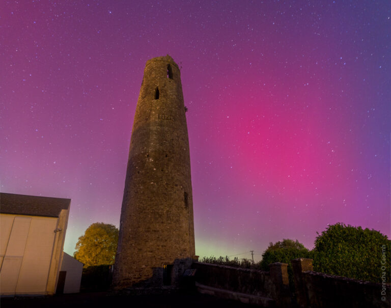 The image depicts a tall, cylindrical stone tower against a vibrant night sky. The sky is illuminated with shades of pink and purple, suggesting the presence of an aurora. Stars are visible throughout the sky, adding to the scene's atmospheric beauty. The tower is marked with the year "1815" and is surrounded by trees and buildings, creating a striking contrast between the historic structure and the colorful celestial display.