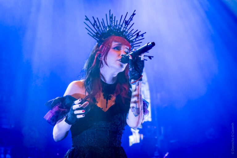 A performer on stage, singing into a microphone. The performer is wearing a striking black outfit with lace details and fingerless gloves. They have a unique headpiece resembling a crown made of spikes. The lighting is dramatic, with blue hues creating a vivid atmosphere. Tattoos are visible on the performer's arm.