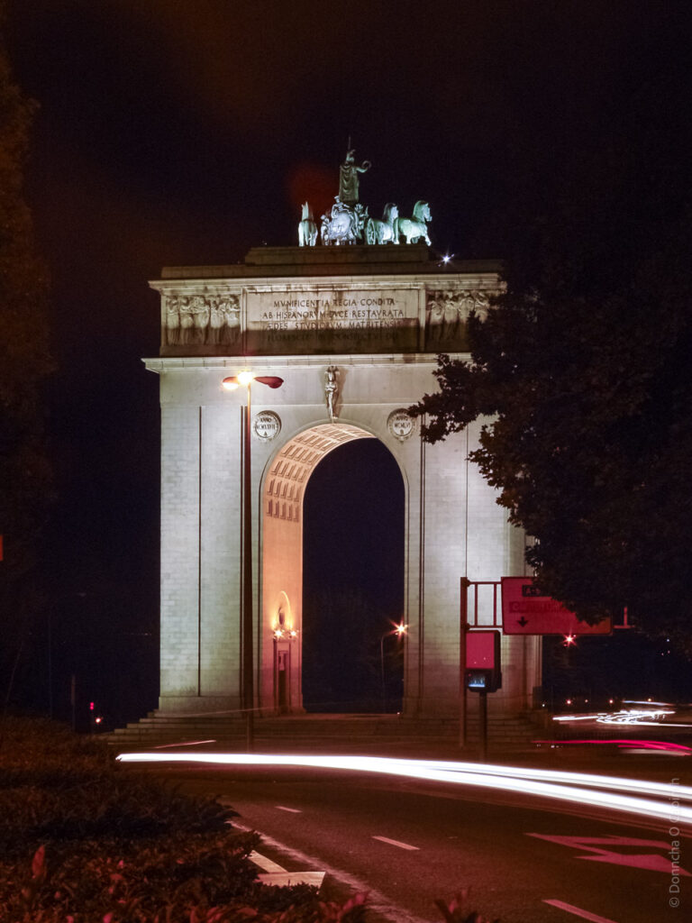 A large triumphal arch illuminated at night. The structure features classical architectural elements, with a prominent archway and decorative sculptures on top, including a chariot with horses. The lighting highlights the details of the arch, and there are light trails from passing vehicles on the road in front of it. Trees are visible on the sides, framing the monument.