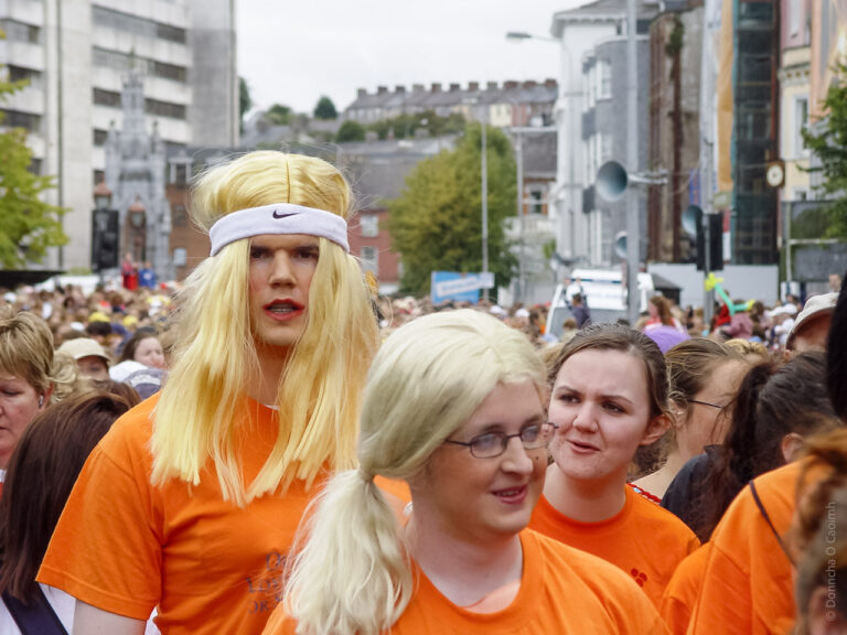 A large crowd of people, with some participants wearing orange shirts. In the foreground, a person with long blonde hair and a white headband stands out. The setting appears to be an outdoor event or parade, with buildings and trees visible in the background. The atmosphere seems lively and festive.