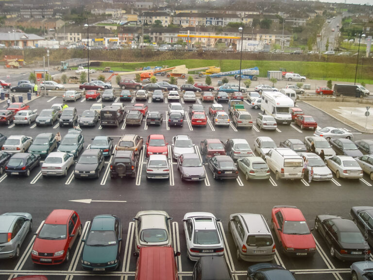 The image shows a busy parking lot filled with numerous cars parked in organized rows. The cars vary in color and type, with some red, silver, and dark-colored vehicles visible. In the background, there is a construction site with machinery and equipment, as well as some residential buildings. The parking spaces are marked with white lines, and there are a few people walking near the vehicles. The scene appears to be taken from an elevated position, providing a wide view of the area.