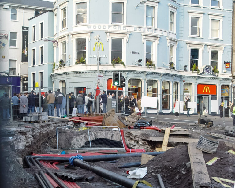 This image shows a similar scene with more focus on a corner building housing a McDonald's restaurant. A crowd of people is gathered outside, and the construction zone is in the foreground with exposed pipes and materials. The building has a sign reading "Woodford, Bourne & Co." above the McDonald's.