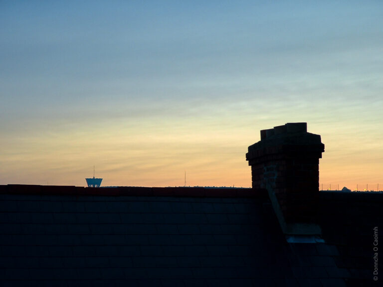 The silhouette of the roof and chimney of a house at sunset while a water reservoir is visible in the background.