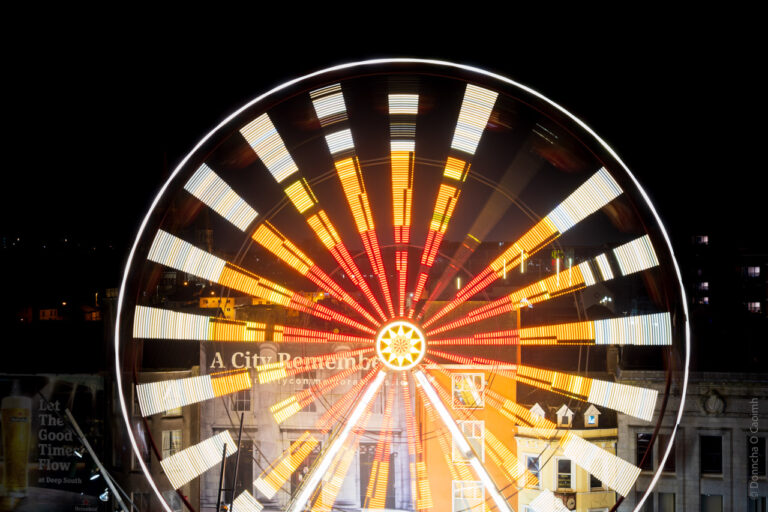 A long exposure photograph of an illuminated Ferris wheel at night, creating a circular pattern of light trails in red, yellow, and white. The wheel's structure appears as a perfect circle against a dark background, with radiating light beams emanating from the center like spokes. The light trails create a mesmerizing starburst effect, while the wheel's frame is visible as a transparent, geometric pattern around the circumference.