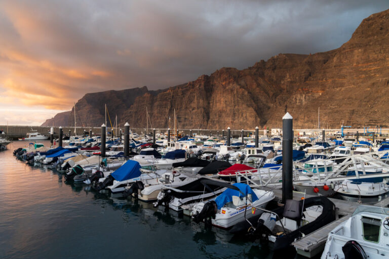 The marina at Los Gigantes, Tenerife, with boats moored against dramatic cliffs at sunset, orange sky reflecting on calm harbour waters.