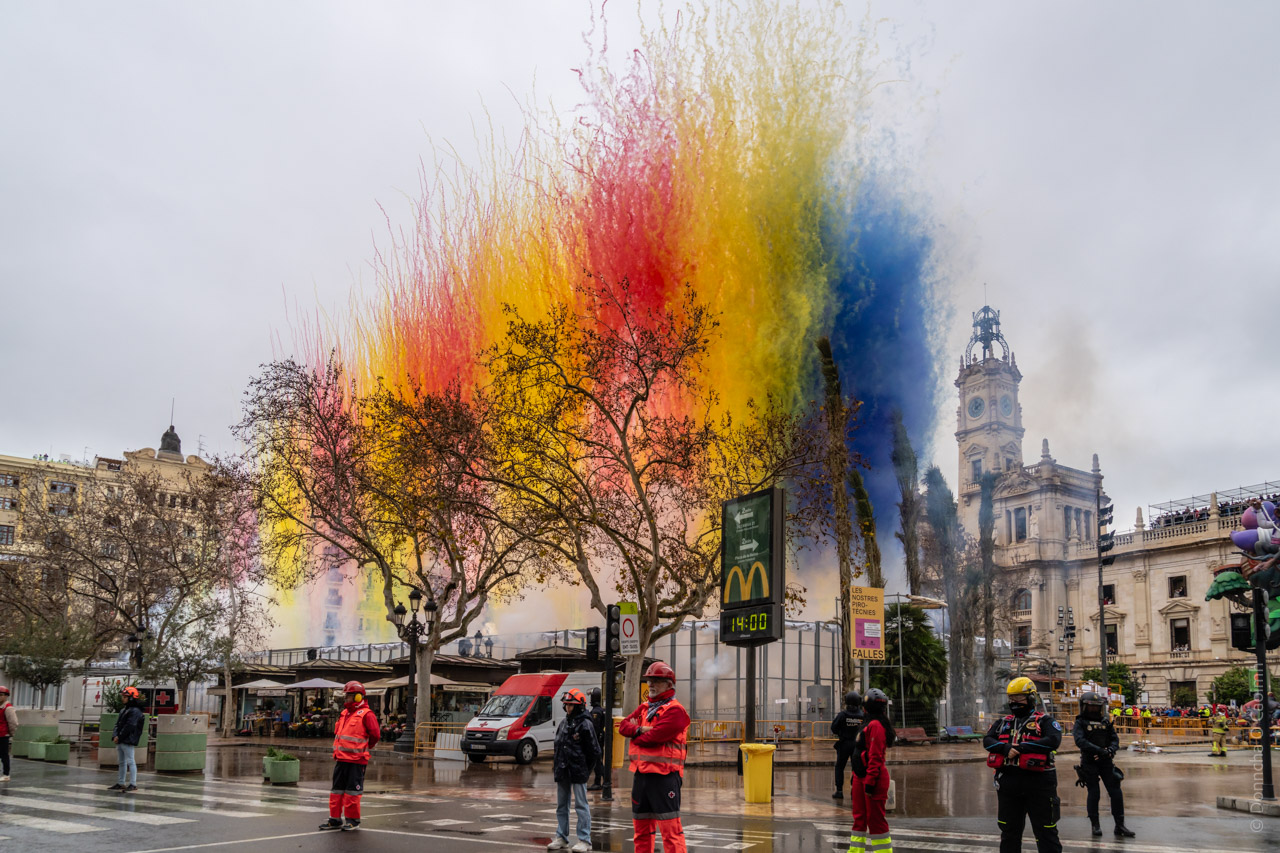 A vibrant display of coloured smoke—yellow, red, and blue—fills the sky during a pyrotechnic event in Valencia, Spain's Town Hall Square. Emergency workers in bright uniforms stand in the foreground as the spectacle unfolds against a backdrop of historic architecture, including the clock tower of the Town Hall. Signs for McDonald's and "FALLES" are visible on the street.