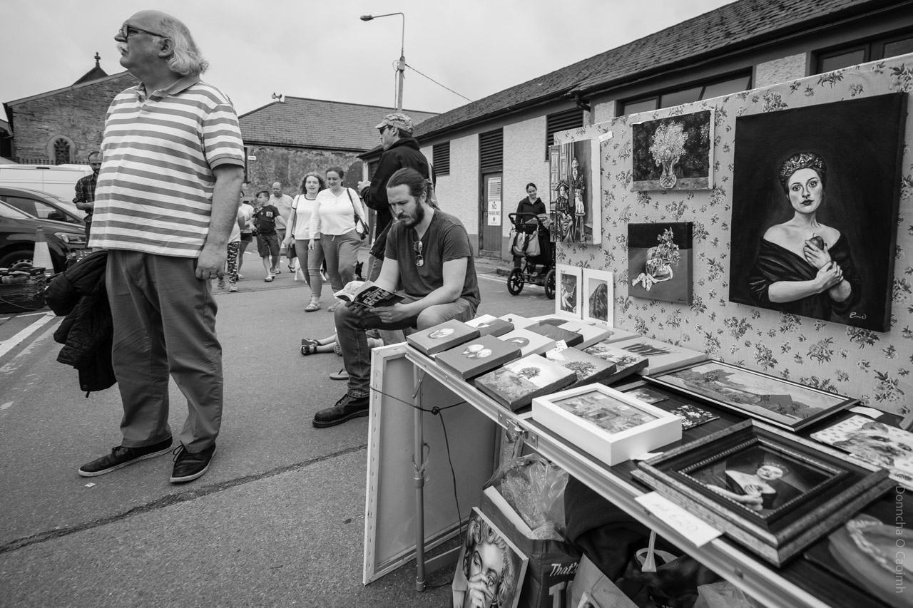 A black and white photo of an outdoor market scene in Skibbereen, Co Cork, Ireland. In the foreground, a table displays framed paintings, books, and art prints, with a floral-patterned backdrop featuring more artwork. Several people are visible: one man stands to the left in a striped shirt, another sits reading a book, and a group of people walk in the background. The setting is lively, with a mix of shoppers and vendors, and the market is held alongside a row of buildings and an old stone church.
