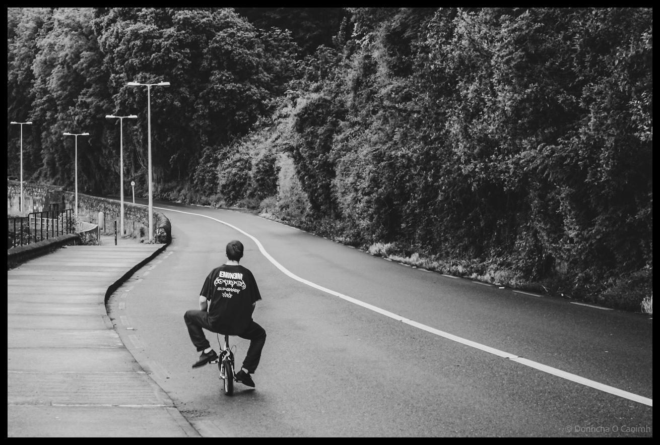 Black and white photograph of young man on small bicycle riding along curved road from Passage West to Monkstown in Co Cork, with footpath, street lamps, dense woodland, and rolling hills in background