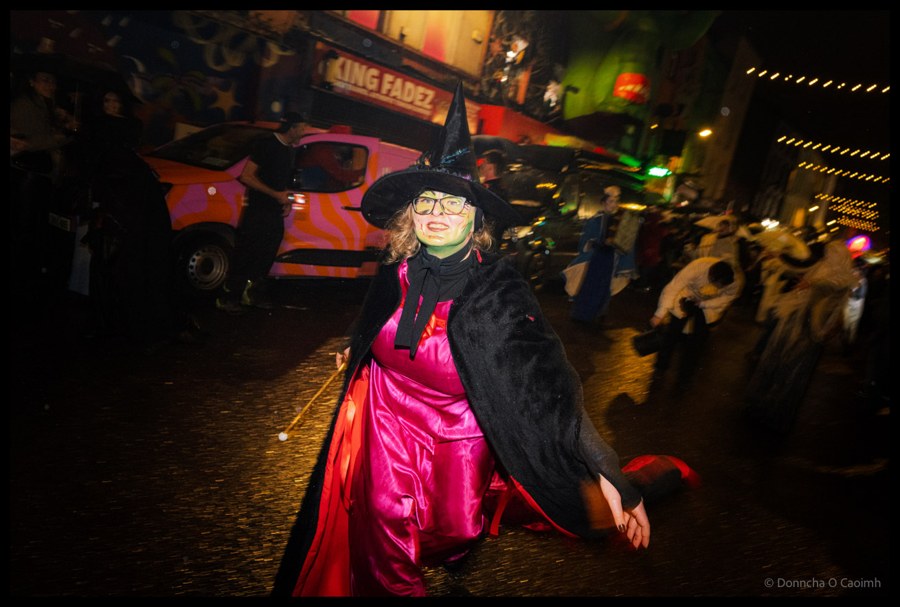 Young woman in elaborate witch costume with black pointed hat, green face paint, hot pink satin dress and black cape poses during Dragon of Shandon parade on North Main Street Cork at night with pink and orange striped van and colourful murals visible behind