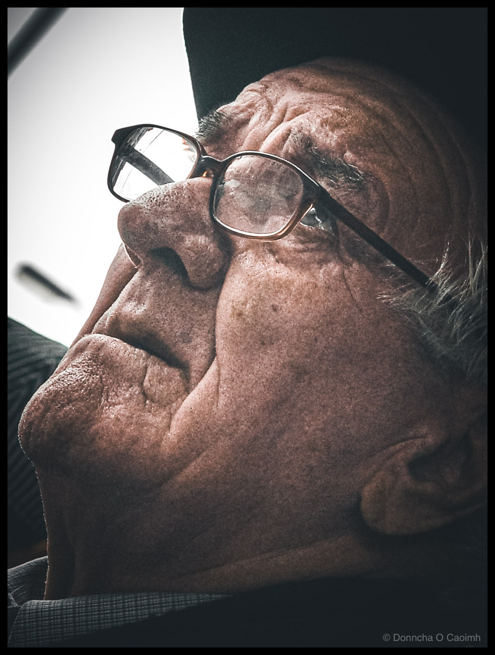 Close-up portrait photograph of an elderly man shot from below as he looks upward, taken outside a cafe on Daunt Square in Cork, Ireland in 2004. The image captures his weathered face in profile, showing deep wrinkles and aged skin texture in rich detail. He wears rectangular glasses with metal frames that catch reflections of buildings and sky, and a dark flat cap. Wisps of grey hair are visible at the back of his head. He wears a checked shirt. The photograph has a dark vignette around the edges with dramatic lighting emphasising the contours of his face.