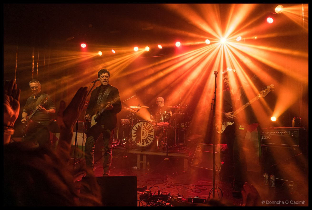 The Stunning performing under intense orange and red starburst stage lighting, with the lead singer playing guitar centre stage, fellow band members on either side, The Stunning drum kit logo visible behind, and audience members' raised hands silhouetted in the foreground.