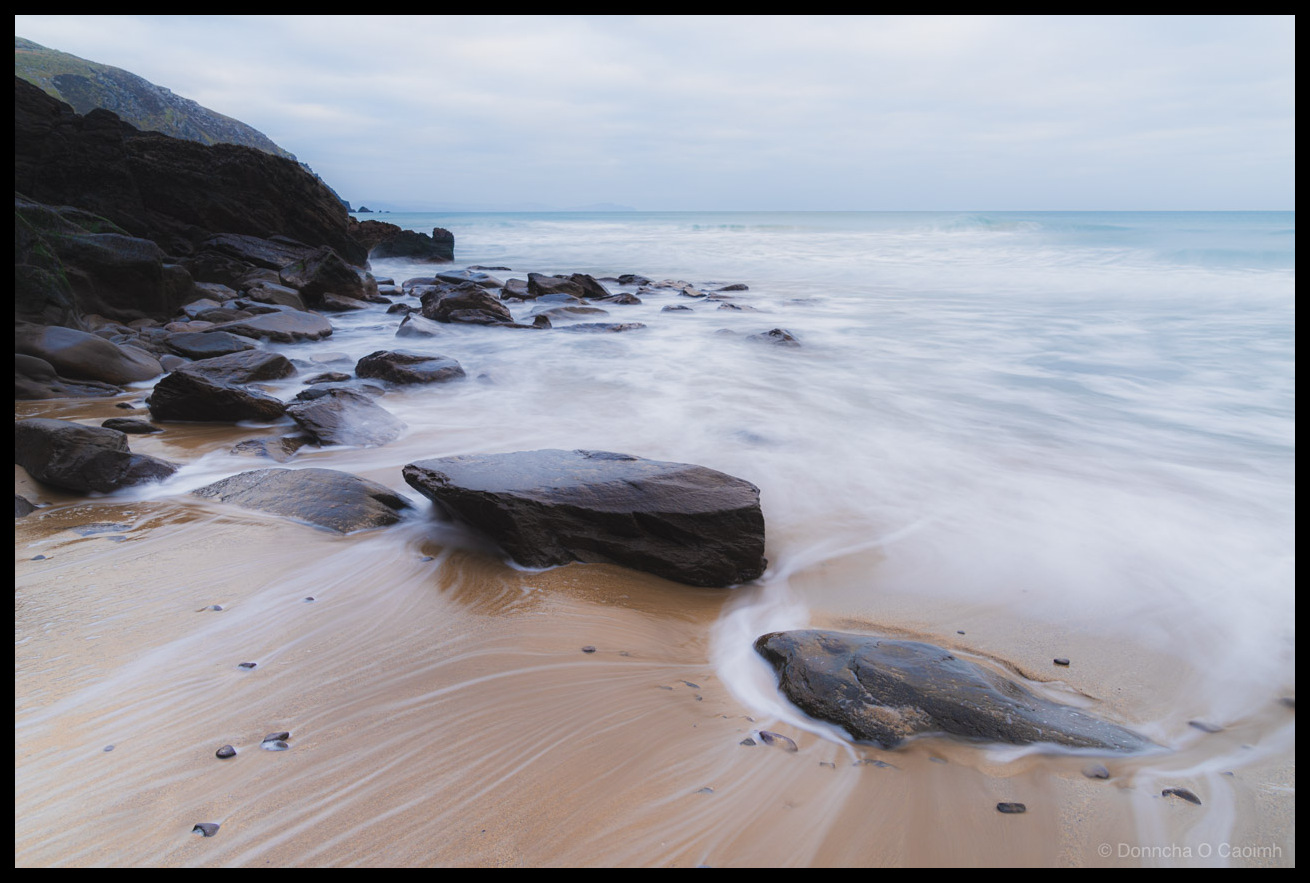 Long exposure photograph of Coumeenoole Beach in County Kerry showing smooth, misty water flowing around dark rocks on golden sand, with dramatic cliffs and mountains under an overcast sky.