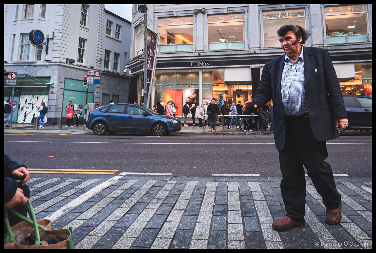 A man in a dark blazer and blue checked shirt gesturing mid-stride on a cobbled pedestrian crossing on St Patrick's Street in Cork, with Penneys behind him, a purple banner reading "Rembrandt" on a lamppost, a parked blue Ford Focus, pedestrians on the pavement, and a sheep-patterned shopping bag held by a seated figure in the bottom-left corner.