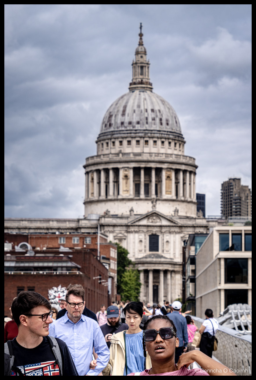 St Paul's Cathedral dome rising above Victorian and modern buildings, viewed from the Millennium Bridge, with a crowd of pedestrians in the foreground under an overcast sky; the nearest figures include a young man in glasses with a dark T-shirt showing a Norwegian flag and partial text "PROGR", a bespectacled man in a pale blue shirt, and a woman in sunglasses and a light blue top.
