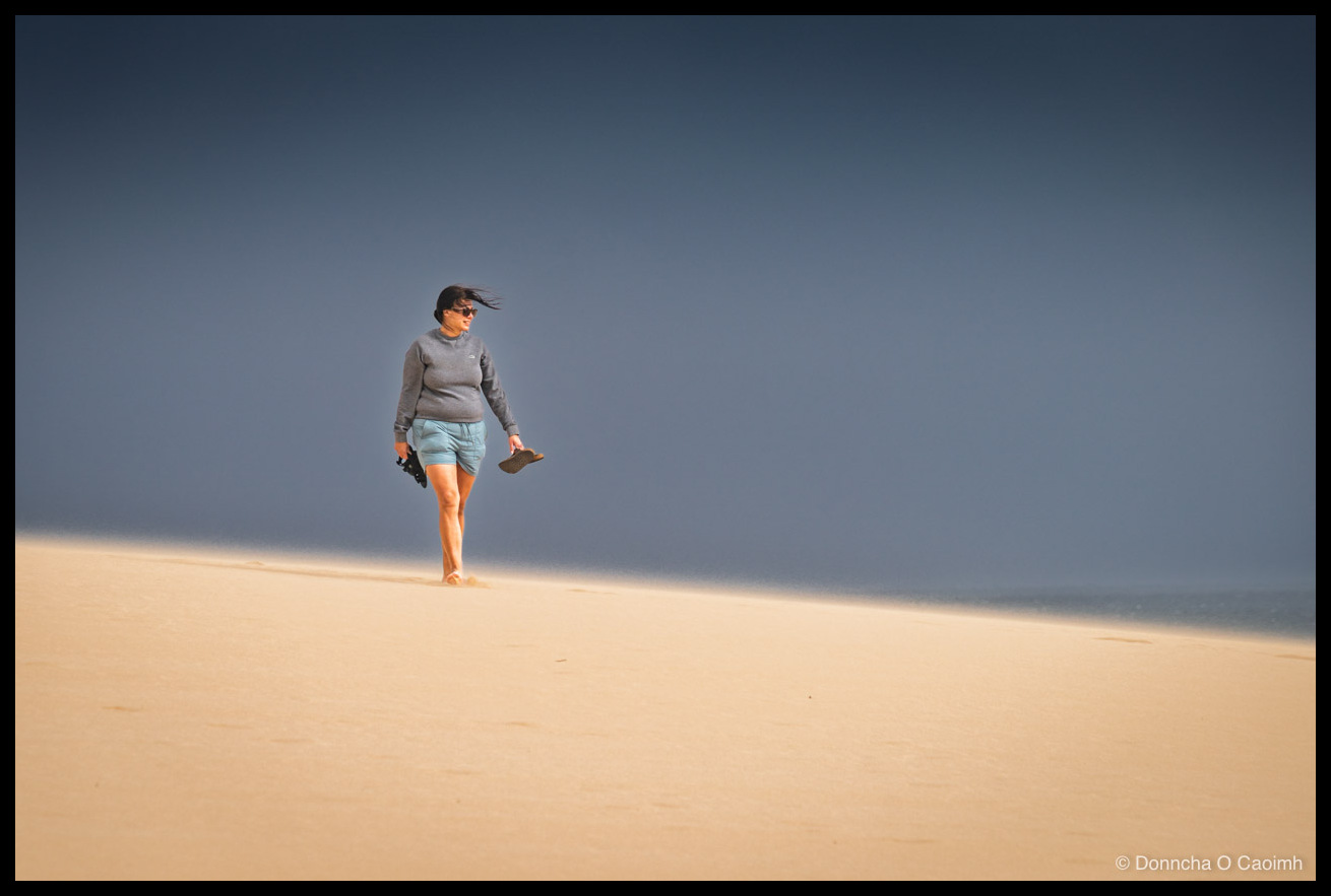 A woman in a grey jumper and pale blue shorts walking barefoot across a sunlit sand dune, sandals in hand, hair blown sideways by the wind, with a moody grey sky above and a thin strip of sea visible on the right.