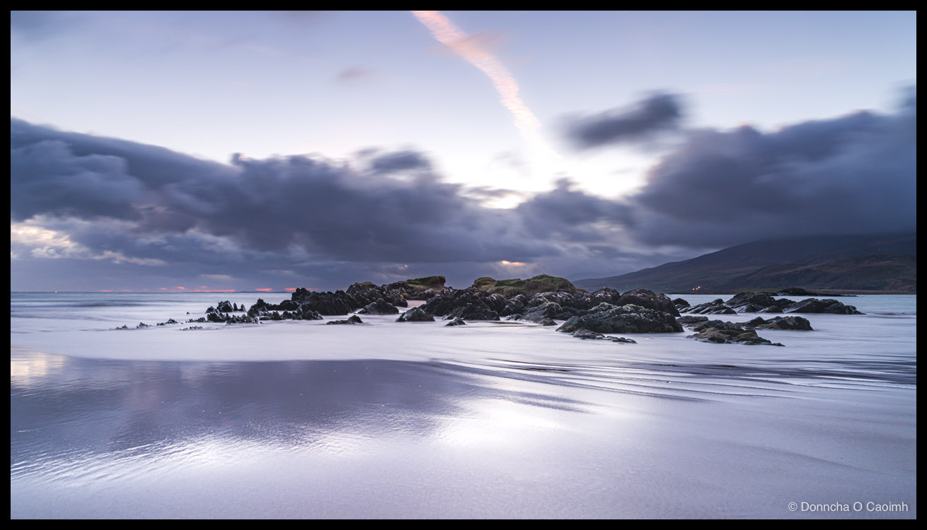 Cappagh Beach in Kerry before dawn, with wet sand reflecting a moody twilight sky, dark rocks in the middle distance, a faint sun glow behind heavy cloud, and a pink contrail streaking diagonally overhead.