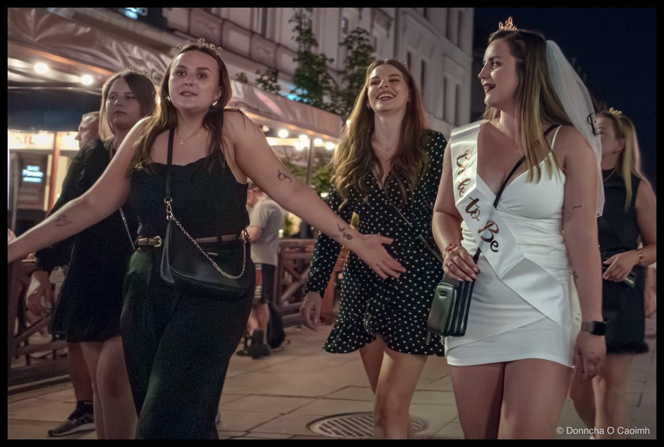 Five young women walking along a Katowice street at night during a hen party; the bride-to-be on the right wears a short white dress, a tiara with veil and a sash reading "Bride to Be"; her friends wear dark dresses, two also wearing small tiaras, with a lit-up restaurant terrace and bystanders behind them.