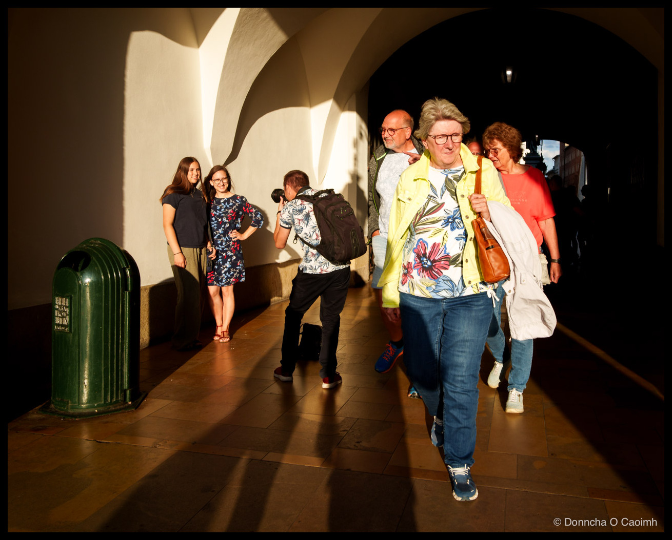 Two young women pose against a sunlit arcade wall in Kraków while a photographer in a floral shirt and black backpack crouches to photograph them, and a group of older tourists walks out of the shadowed passage towards the camera, led by a woman in a bright yellow jacket over a floral top.