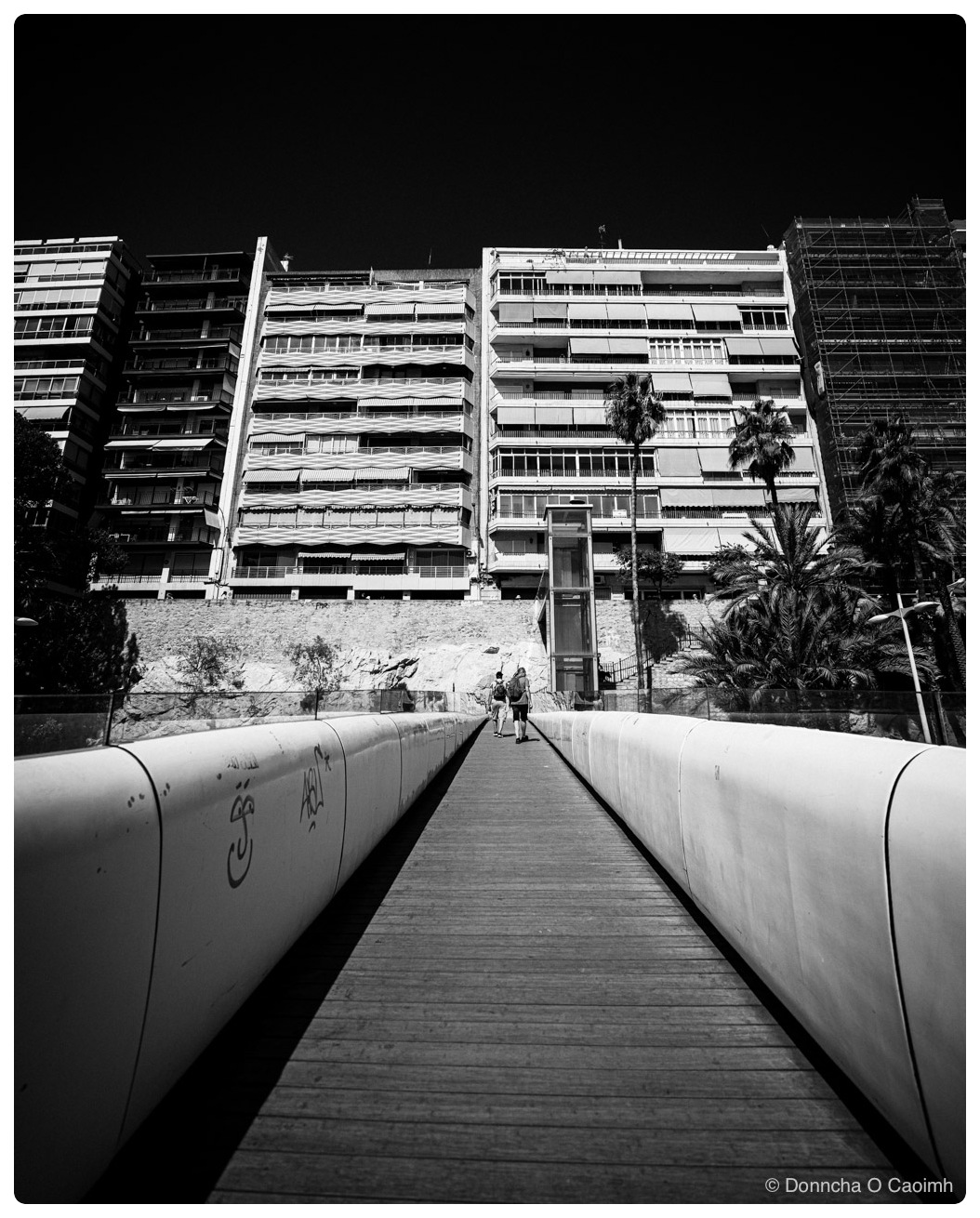 Black-and-white view along a narrow wooden footbridge with tall, curved white side walls, one marked with two graffiti tags, leading towards a sunlit rock wall where two people with backpacks are walking; above the wall stand several apartment blocks with balconies and lowered striped awnings, one of them covered in scaffolding, with a glass lift tower and steps at the foot of the wall, palm trees on the right and a nearly black sky overhead.