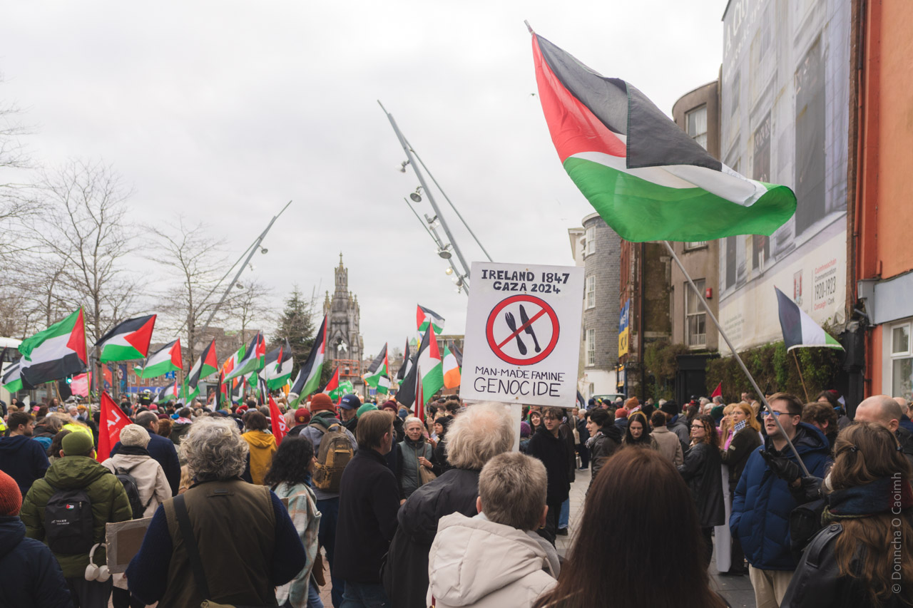Large pro-Palestinian protest crowd in Cork city center with Palestinian flags and sign reading "IRELAND 1847 GAZA 2024 MAN-MADE FAMINE GENOCIDE".