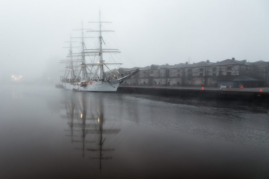 A white sailing ship moored at a quay on a foggy morning. It reflects in the water of the river.