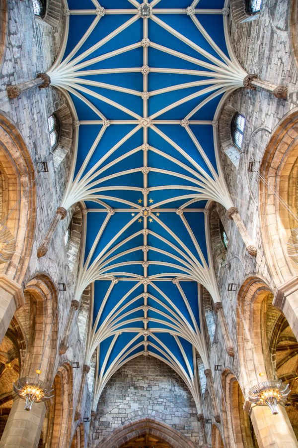 Looking up at the ornate vaulted ceiling of St. Giles Cathedral in Edinburgh, showing a striking blue plaster vault with cream-coloured ribbed arches, decorative bosses, and a cluster of gold stars at the centre, framed by grey stone walls and arched clerestory windows.
