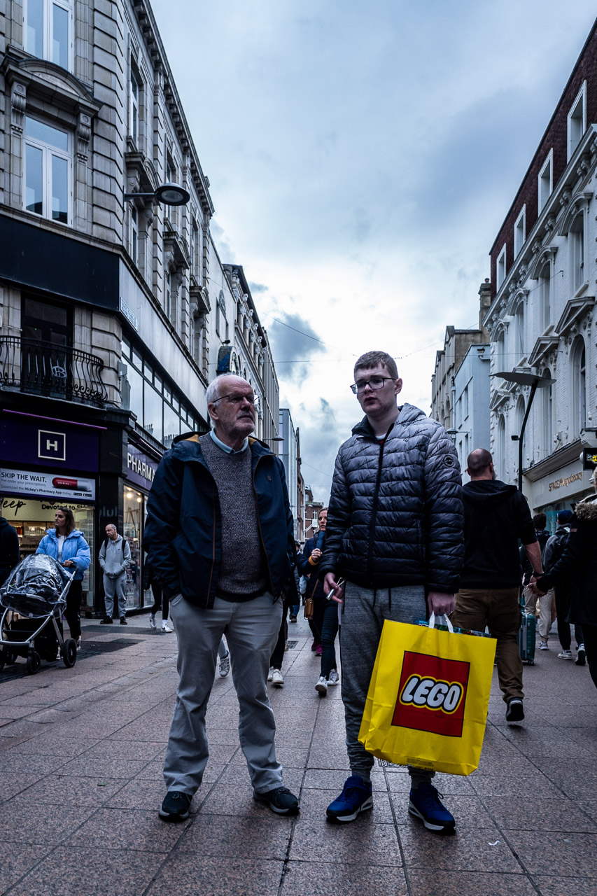 An older man and younger man standing in the middle of a crowded street with a LEGO bag