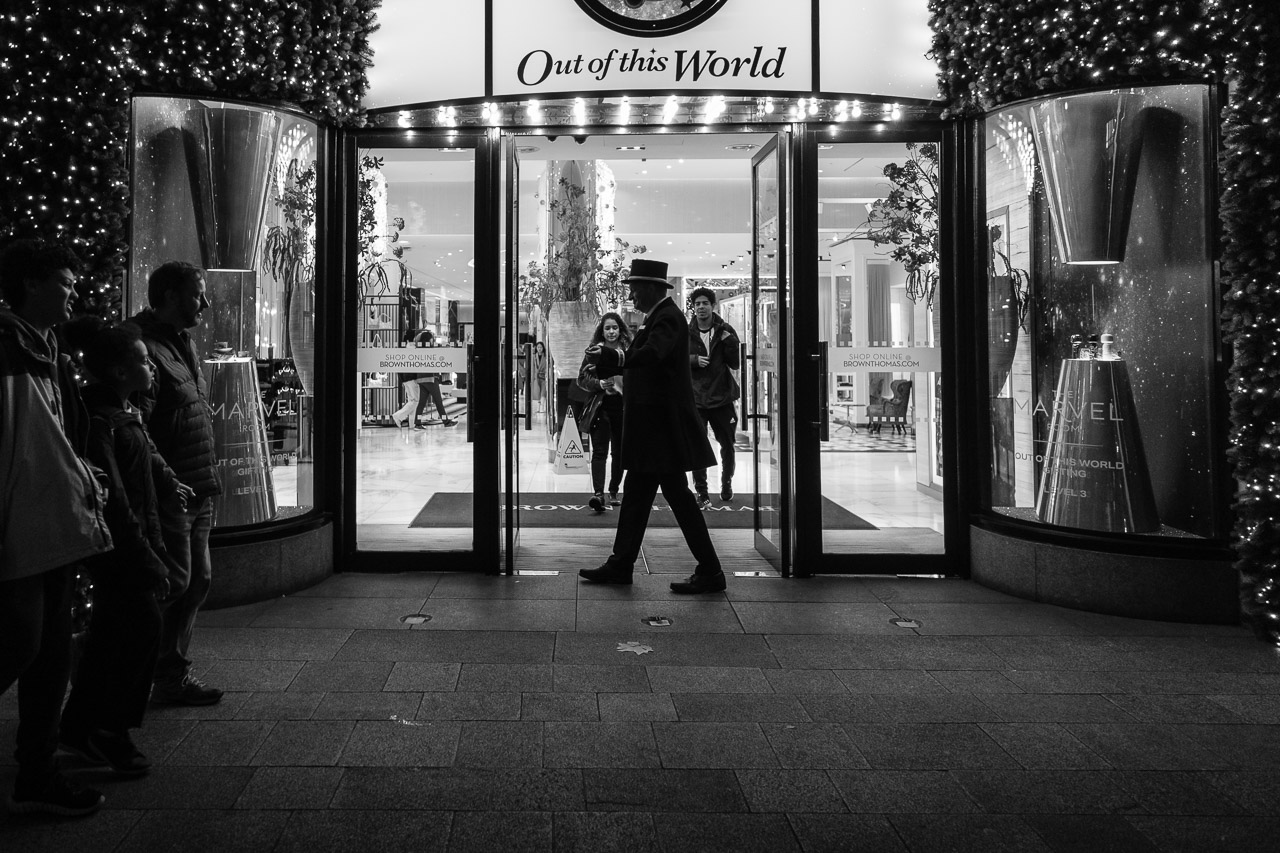 People walking past a shop while the doorman is framed in the front door.
The text "out of this world" appears above the door 