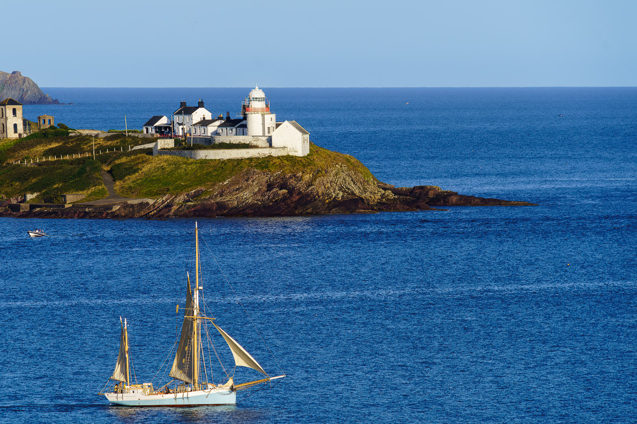 A sailing boat sails past Roches Point Lighthouse at the mouth of Cork Harbour. The boat has a blue hull and the sails are, I think, half raised.