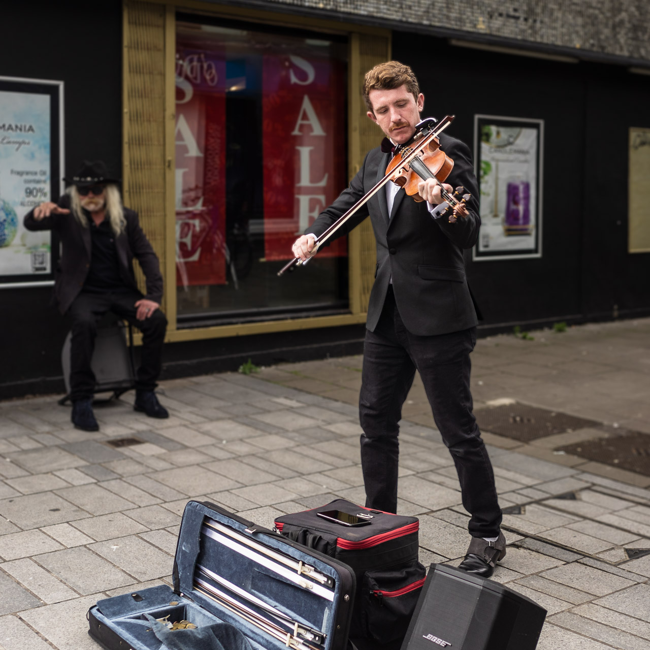 A man playing a violin on the street, with an older man sitting in the background with his arm raised, enjoying the music