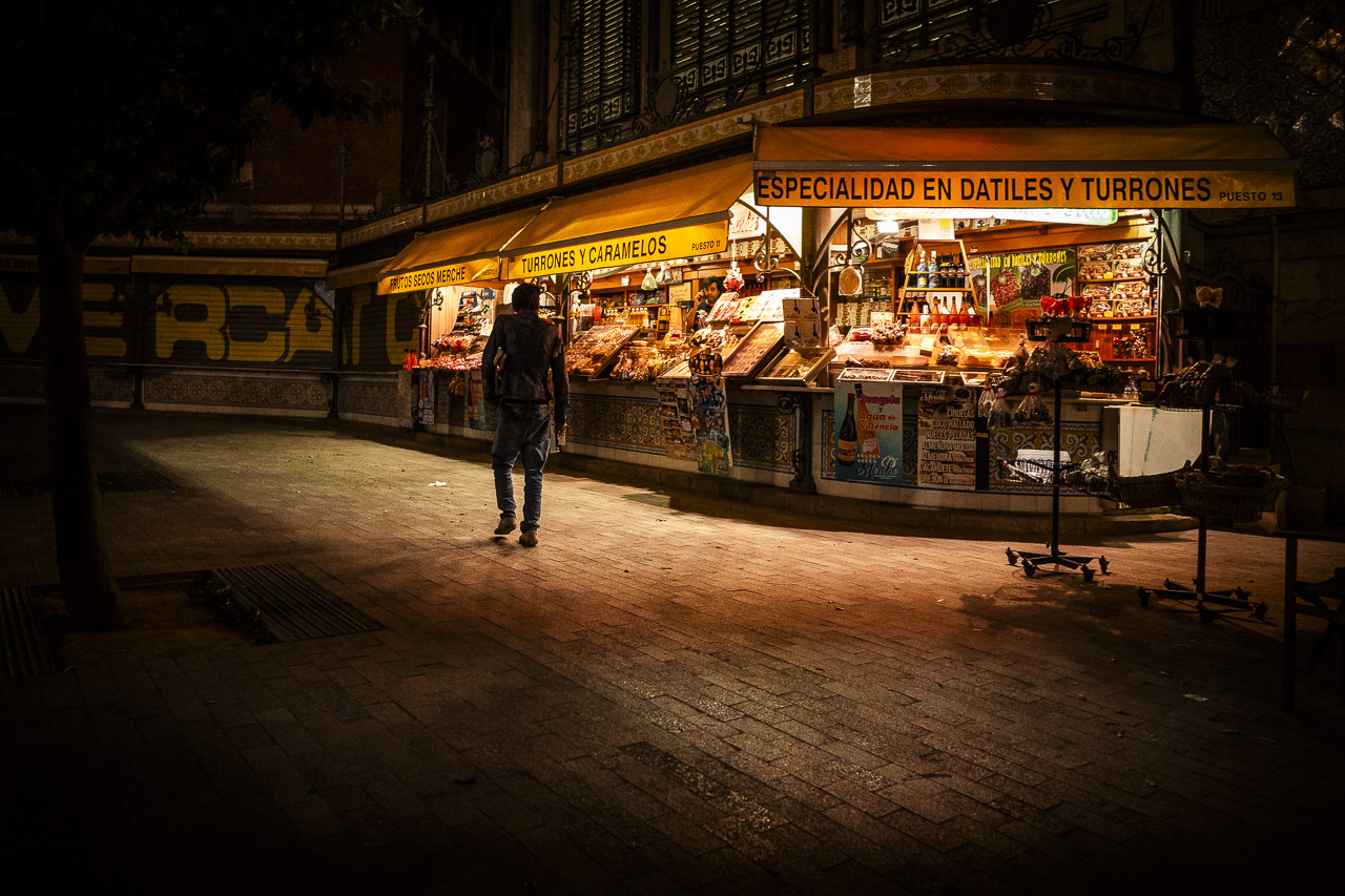 A man walks by a stall selling bread and other items at night. The ground around the stall is lit up bur darkness is all around. Graffiti in the background says, "E RCA".