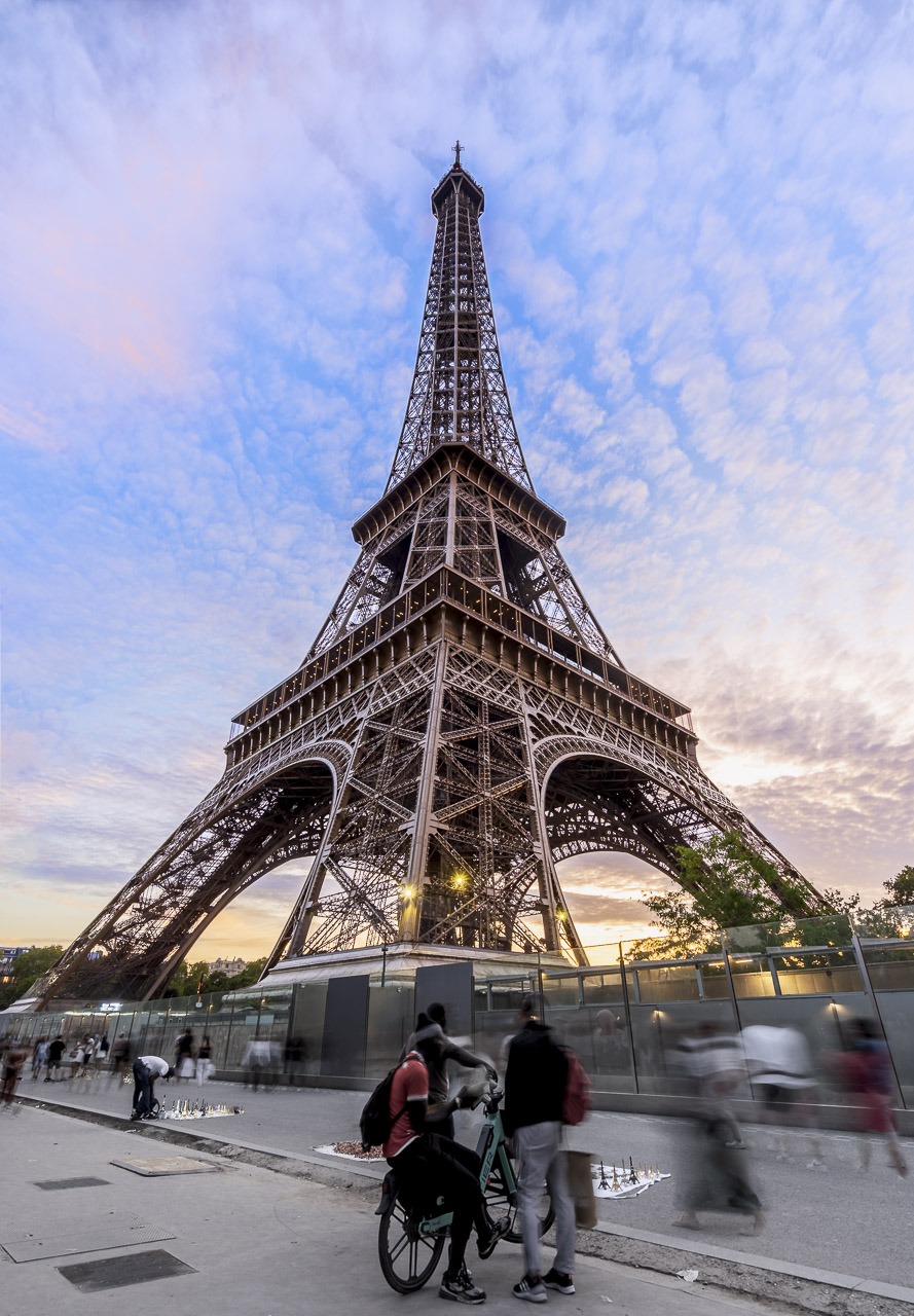 The Eiffel Tower looms over the footpath outside the barrier surrounding the tower. People walking past are a blur while a few stationary groups and a man arranging his plastic Eiffel Towers on the ground.