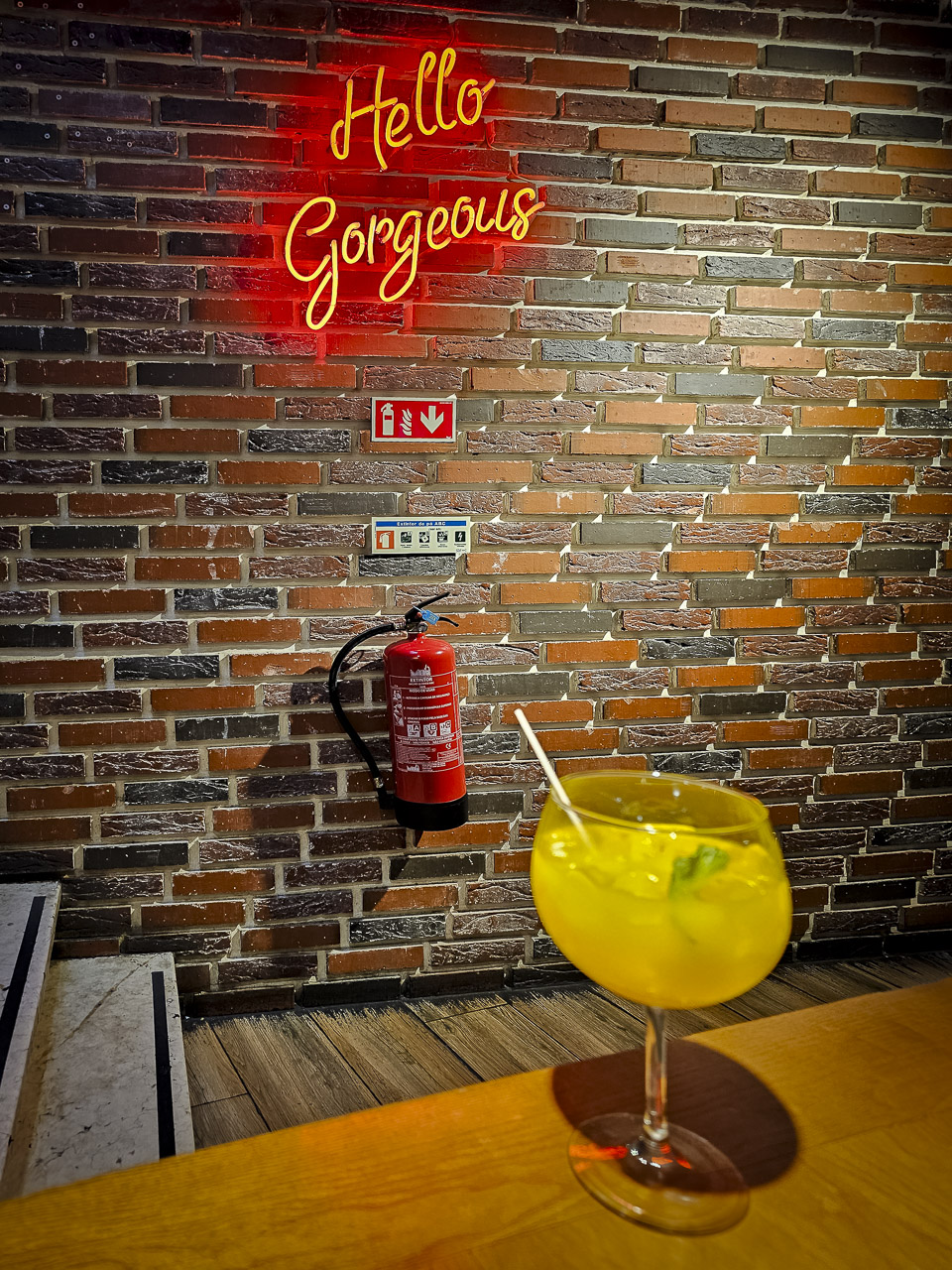 A fire extinguisher on a brick effect wall with a glowing neon sign above it saying "Hello Gorgeous".

A yellow drink in a glass sits on a table in the foreground. The steps of a stairs lead off to the left.