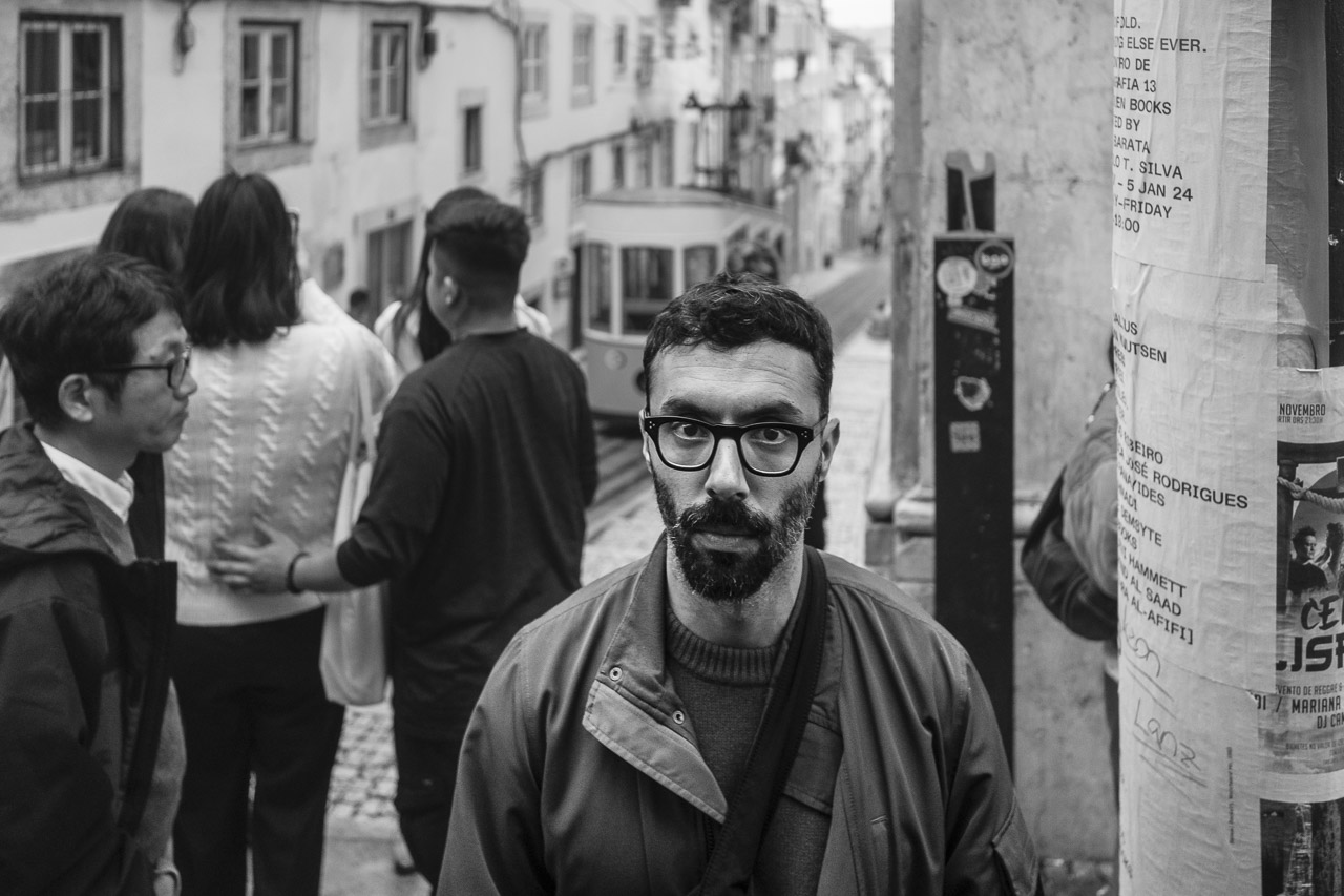 A man looks directly into the camera lens while around him people walk around or stand in the background looking at a tram coming up the road.