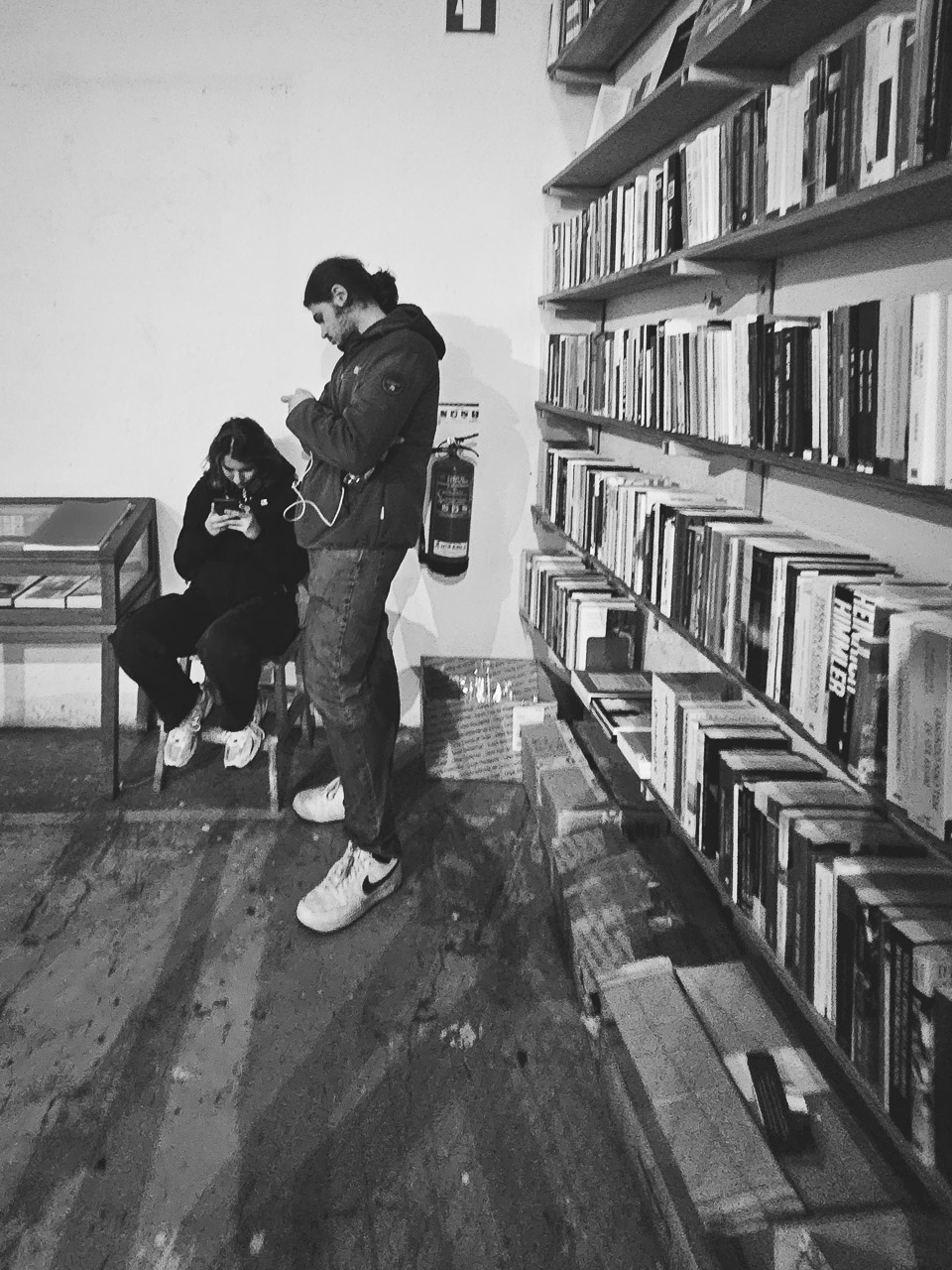 Two people looking at their phones in a bookshop. Shelves of books line the wall.