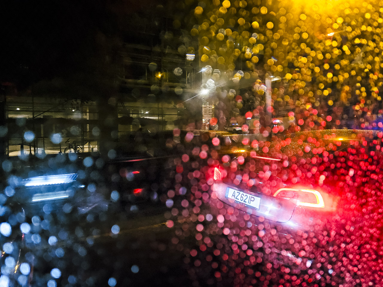 Pictured through a window laden with drops. Warm red and orange lights from street lighting and the rear brakes of a Tesla win against the cold, almost blue headlights of a passing car.
A bike can be seen in the background.