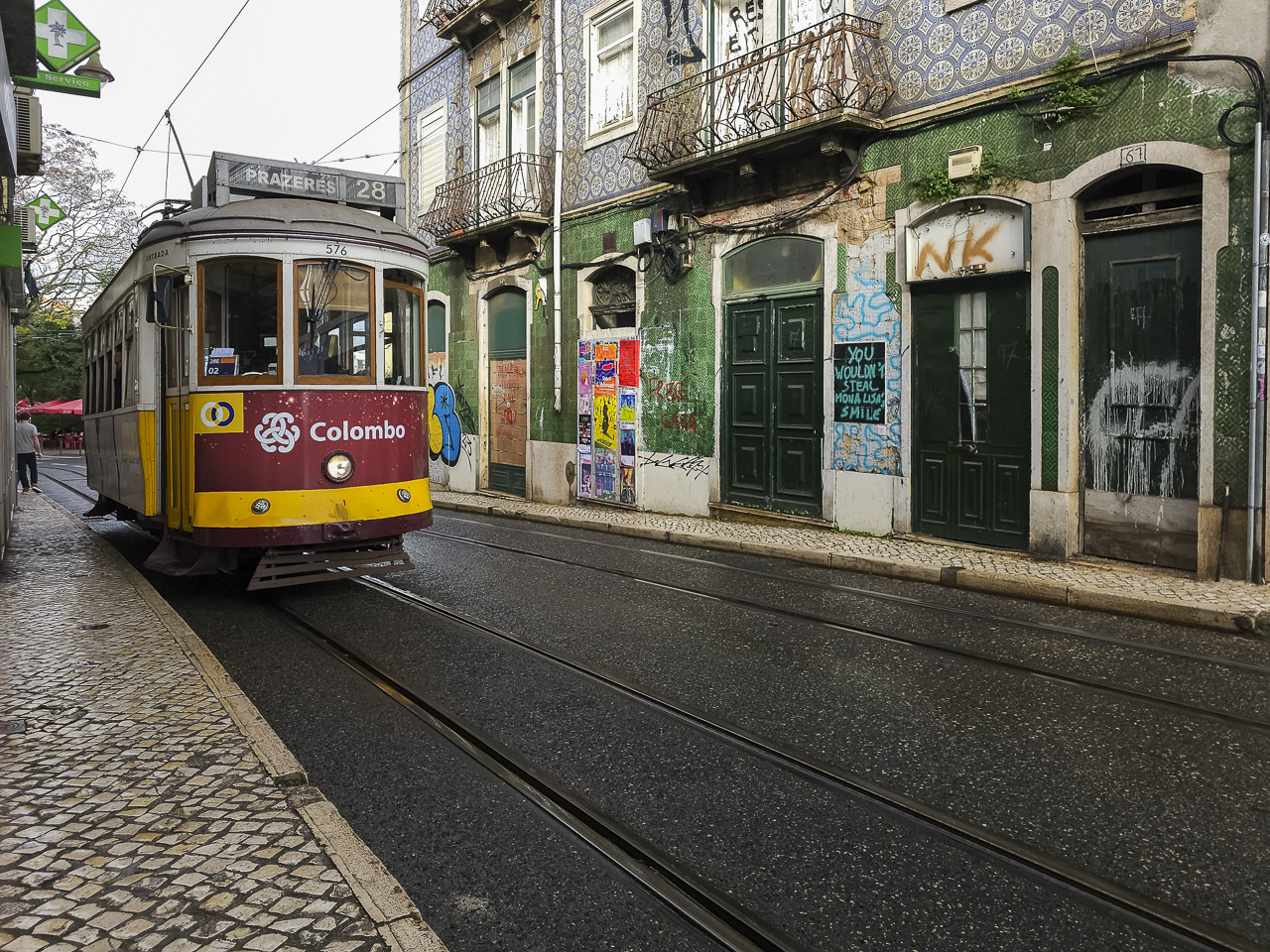 A tram on line 28 in Lisbon rumbles up the street along Rua da Graça. Old houses line the opposite side of the street with graffiti on the walls.