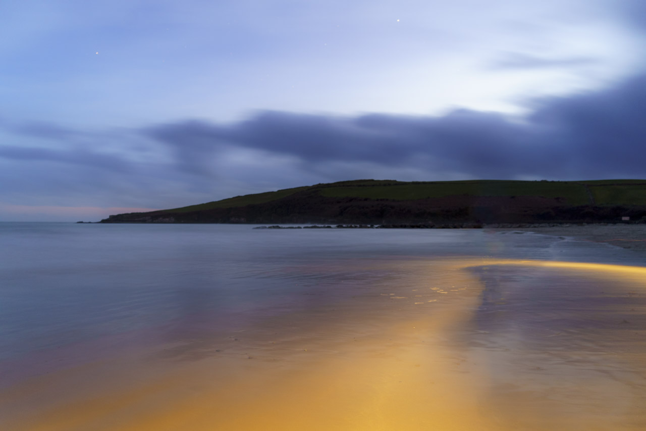 A long exposure photo of a beach, with a glowing luminescence in the water and clouds in the sky.
A hint of the setting sun can be seen at the horizon.