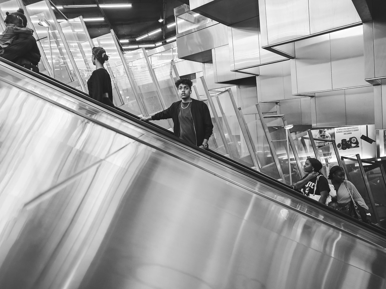 People going up an escalator. Smooth polished metal surfaces. On the escalator are a:
1. parent/guardian holding a child.
2. a woman looking away.
3. a man looking towards the camera. He has a gold chain around his neck.
4. two women near the bottom of the photo.