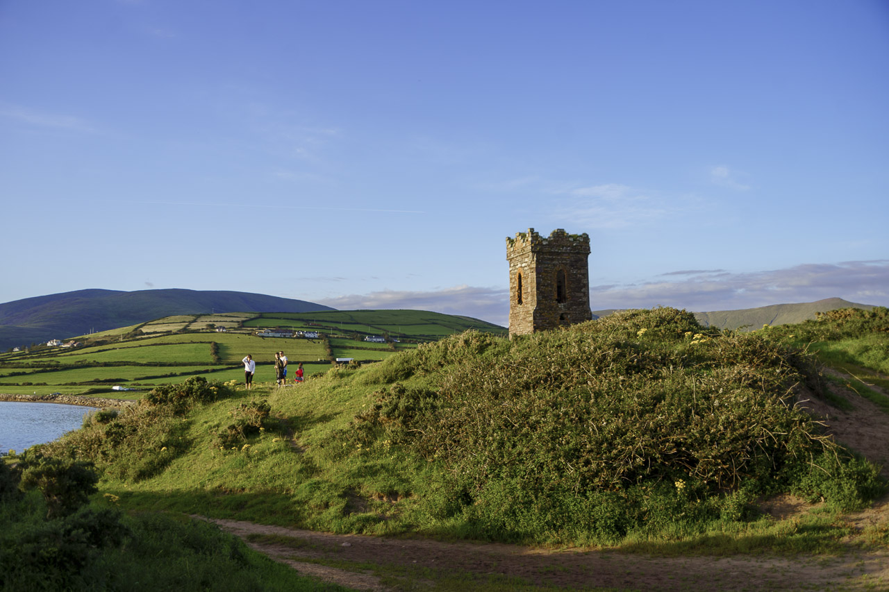 A small castle, or folly, sits on a small grassy hill. People are visible nearby, and green fields stretch off in the distance.