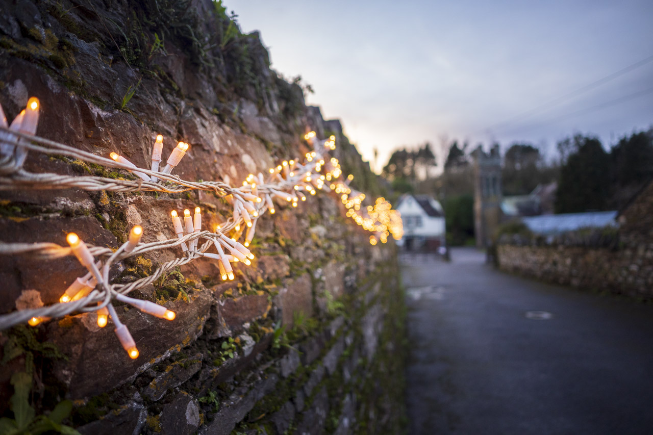 Christmas lights line a wall on a road heading to a church and house in the background.
