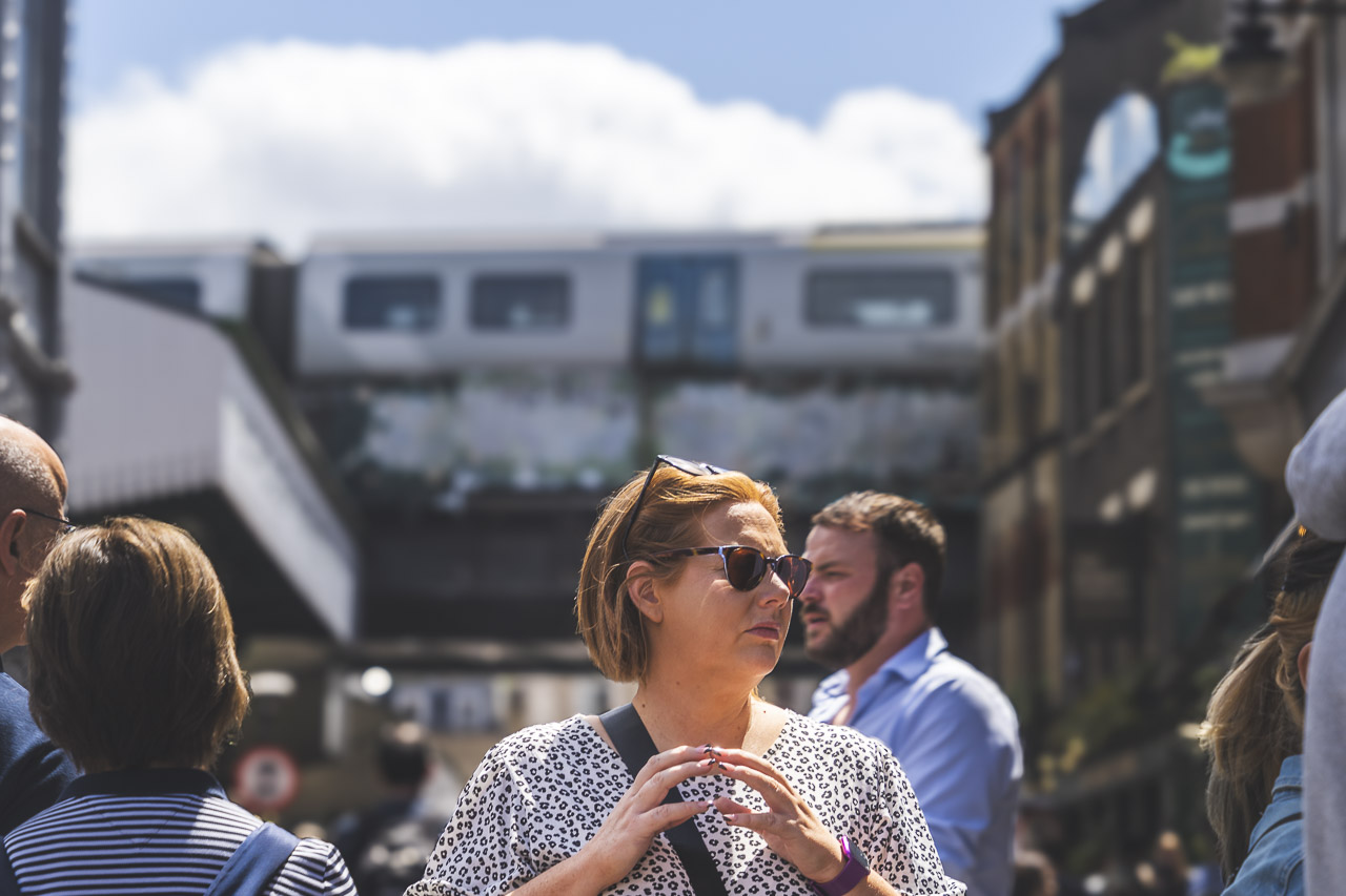 A woman with her fingers steepled looks to her side at something off camera.
Lots of people are walking about on a sunny day, and a train is seen in the background going past on a bridge.