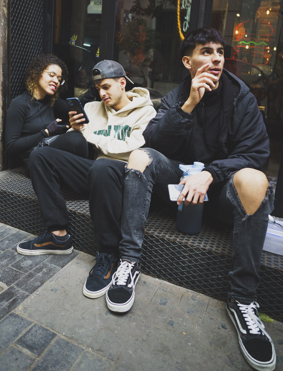 A group of young people sitting on a window sill down a side street.
A woman and two men. They each have phones and the man on the right is speaking to someone outside the frame.