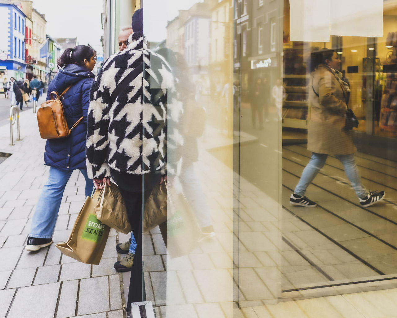 A woman wearing a black and white patterned jacket leans against the window of a clothes shop. Other people walk in and a male head can just be seen over her shoulder.